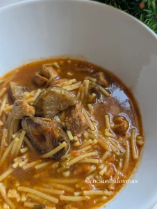 Cazuela de fideuá de carne con alcachofas y verduras en un caldo espeso de color dorado.