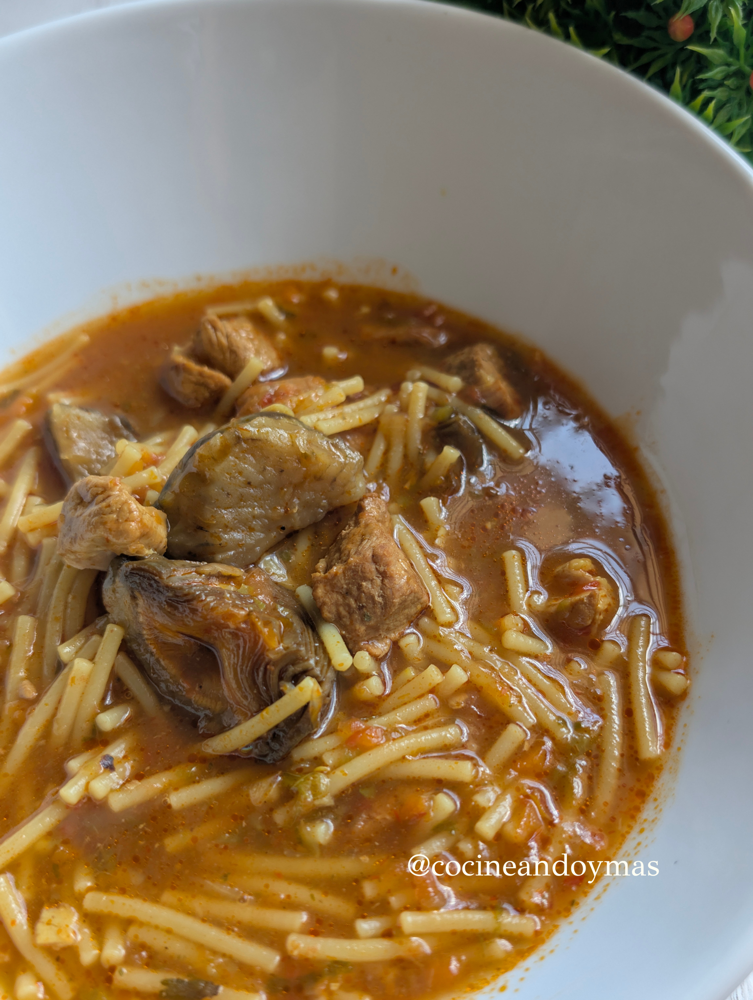 Cazuela de fideuá de carne con alcachofas y verduras en un caldo espeso de color dorado.