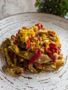 Revuelto de huevos muy jugoso con setas salteadas, tiras de pimiento del piquillo rojo y trocitos de jamón en una sartén negra.