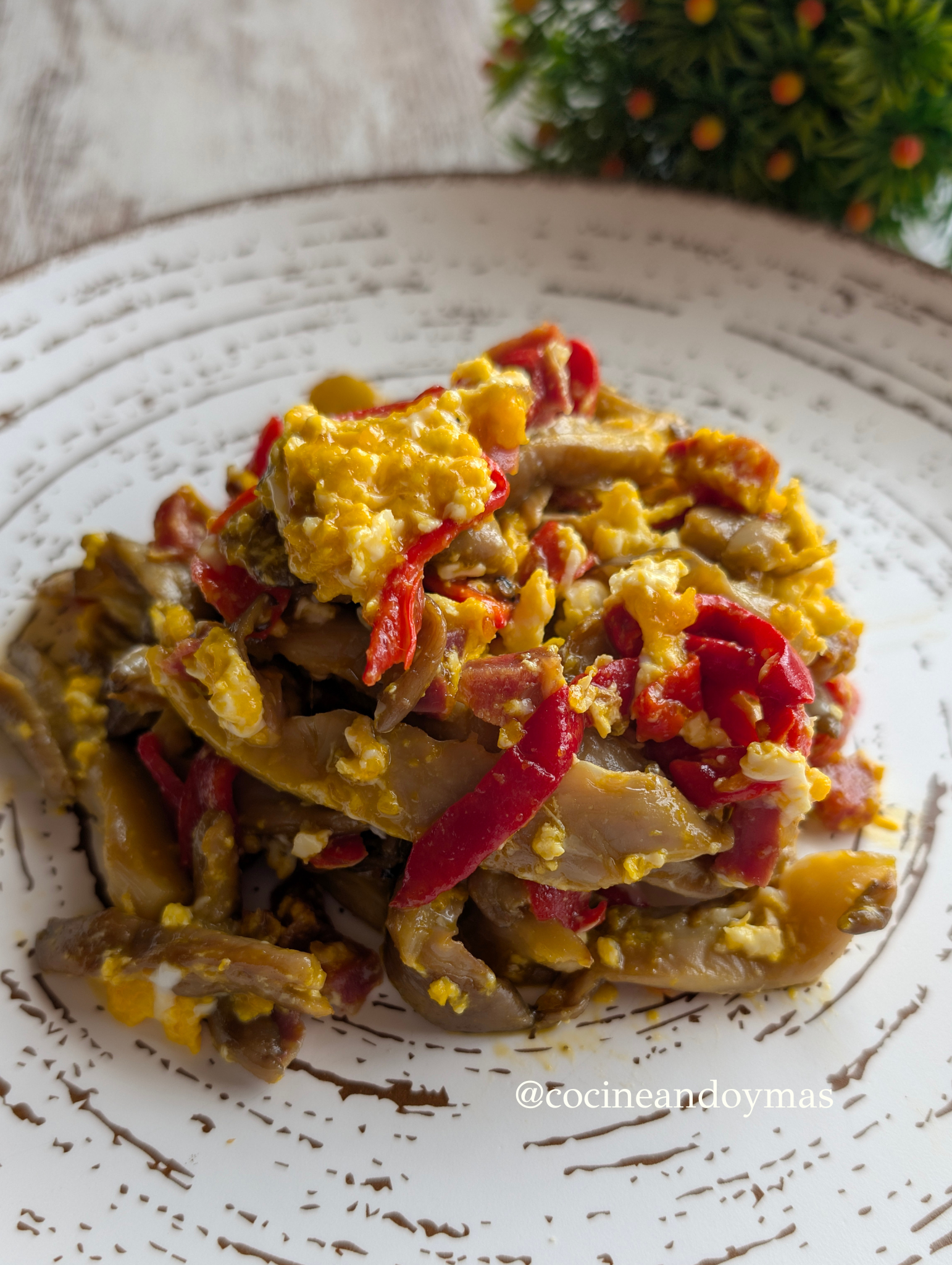 Revuelto de huevos muy jugoso con setas salteadas, tiras de pimiento del piquillo rojo y trocitos de jamón en una sartén negra.