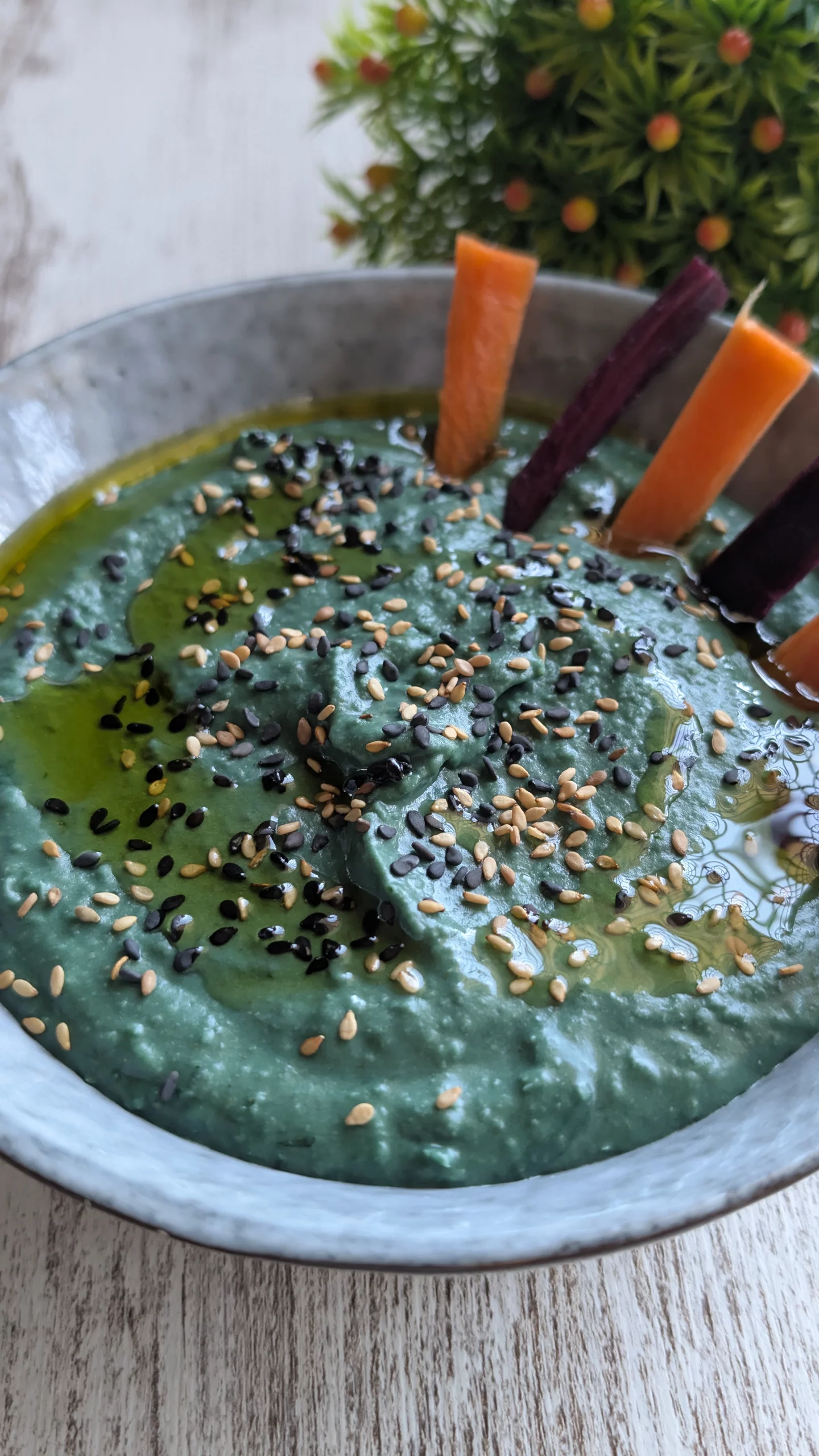 Bol de hummus de color verde intenso con espirulina, decorado con semillas de sésamo y aceite de oliva, servido con crudités de verduras.