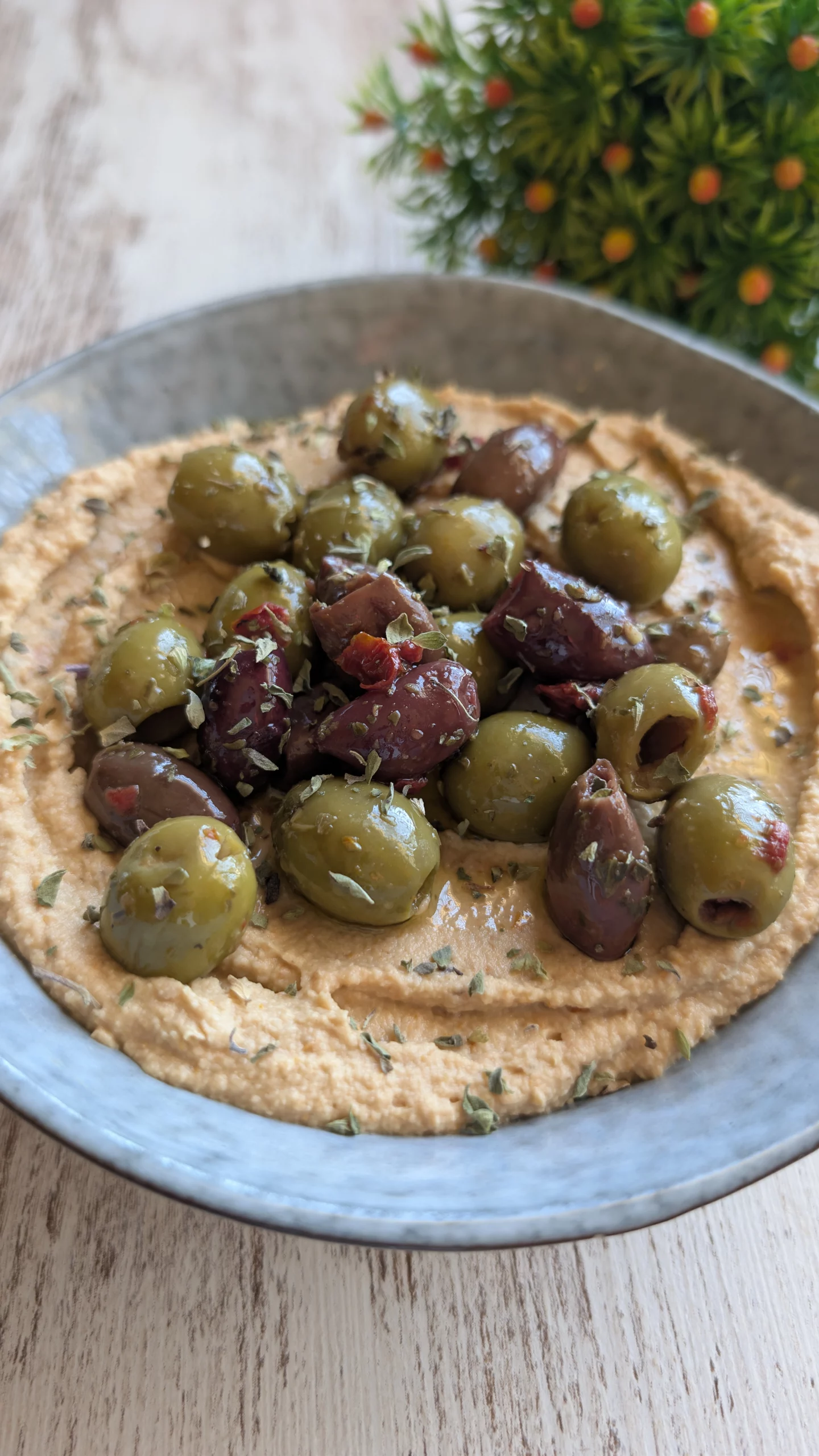 Bol de hummus de color anaranjado con tomates secos, decorado con aceitunas, orégano y un chorrito de aceite de oliva, acompañado de bastones de zanahoria.