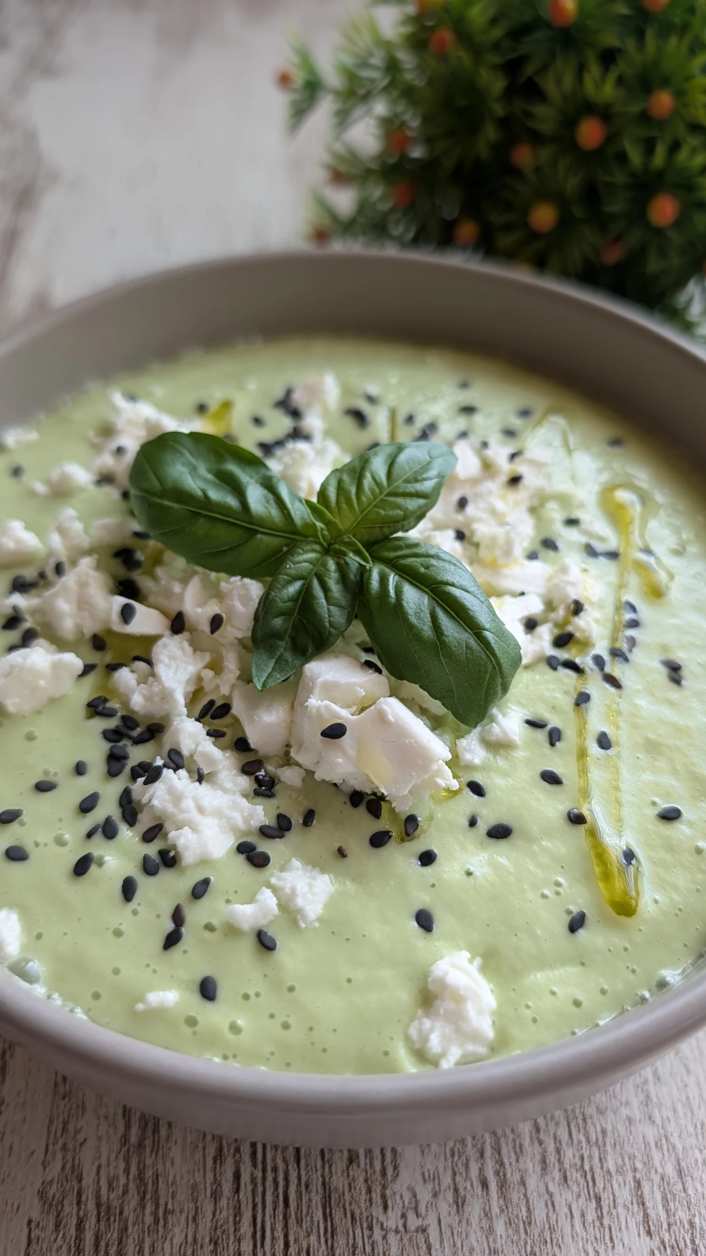 Cuenco blanco con crema fría de color verde claro, decorada con dados de queso feta, semillas de sésamo negro y hojas de albahaca fresca.