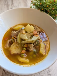 Cazuela tradicional de barro con guiso de patatas, trozos de solomillo de cerdo, alcachofas, setas, guisantes y un caldo espeso de color rojizo.