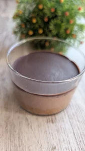 Vasito individual de cristal con capa de almendra en la base, crema de yogur y cacao en el centro y cobertura de chocolate negro crujiente quebrada por una cuchara.