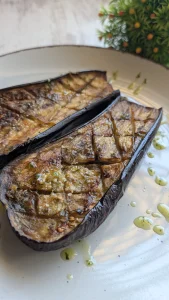 Mitades de berenjena asada al horno con rejilla en la pulpa, doradas y cubiertas de salsa verde de perejil, ajo y aceite de oliva virgen extra.