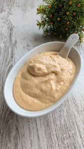 Tarro de cristal con mayonesa de kimchi cremosa de tono anaranjado, servida con una cuchara de madera y acompañada de trozos de kimchi fresco.